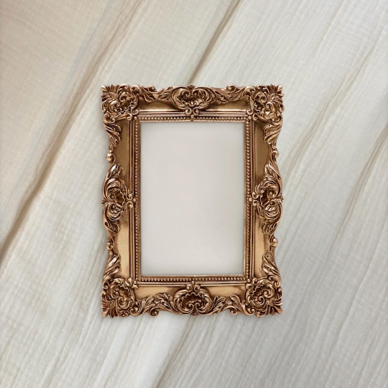 May include: Ornate gold-colored picture frame with a rectangular opening. The frame features detailed scrollwork and decorative elements. The frame is set against a neutral, textured background.
