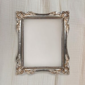 May include: Ornate silver picture frame with detailed floral and scrollwork designs. The frame has a rectangular opening and a slightly curved profile. The frame is set against a white background.