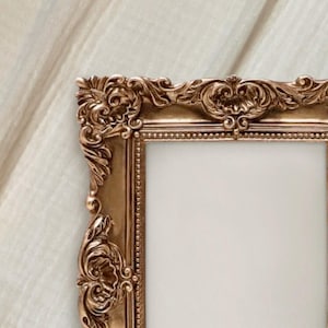 May include: Ornate, gold-colored picture frame with detailed floral and scrollwork designs. The frame has a rectangular opening and is set against a soft, off-white background. The frame's intricate carvings add a touch of elegance.