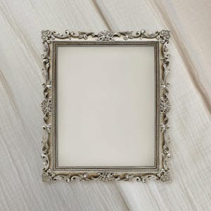 May include: A rectangular silver-colored picture frame with ornate floral and scroll detailing. The frame has a cream-colored interior and is set against a white, textured background. The frame's design is symmetrical and classic.