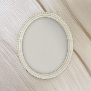 May include: An oval, off-white picture frame with a beaded edge. The frame is empty, with a blank, light-colored interior. The frame is set against a soft, textured, off-white fabric background.