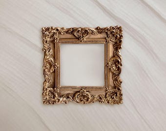 May include: A square, ornate gold picture frame with intricate floral and scroll detailing. The frame has a blank, white center, and is set against a soft, textured background.