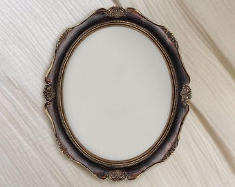 Könnte beinhalten: Ovaler, verzierter Bilderrahmen mit dunkelbraunem Finish und goldenen Verzierungen. Der Rahmen hat dekorative Schnörkel an den Ecken und Kanten. Die Mitte ist ein leerer, weißer Oval, bereit für ein Foto oder Kunstwerk.