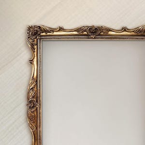 May include: A rectangular picture frame with an ornate, antique gold finish. The frame features intricate, raised scrollwork and leaf-like carvings along the edges, with a beaded inner border surrounding the empty, light-colored center space.