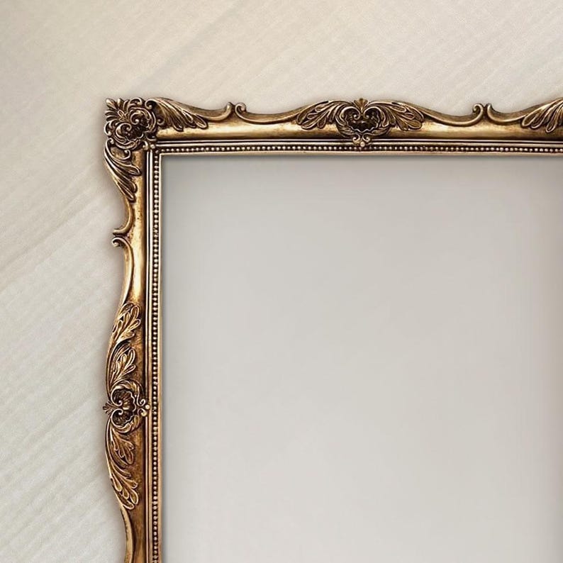 May include: A rectangular picture frame with an ornate, antique gold finish. The frame features intricate, raised scrollwork and leaf-like carvings along the edges, with a beaded inner border surrounding the empty, light-colored center space.