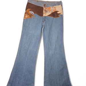 Op de afbeelding: Lichtblauwe denim flared jeans met een decoratief paneel in de taille. Het paneel heeft bruin suède en bloemen en abstracte ontwerpen in oranje en crème. De jeans hebben een klassiek ontwerp met vijf zakken.