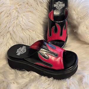 Puede incluir: Sandalias de plataforma negras con detalles de llamas rojas. Las sandalias tienen el logo de Harley-Davidson en la plantilla. El diseño de punta abierta presenta una suela gruesa y un estilo sin cordones.