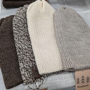 Peut inclure: Trois bonnets en tricot de couleur marron, blanc et beige. Les bonnets sont faits d'un fil doux et texturé. Les bonnets sont disposés en rangée sur une surface blanche. L'étiquette en bas à droite indique "PineKnit".