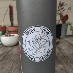 K&ouml;nnte beinhalten: Eine graue Wasserflasche mit einem wei&szlig;en Aufkleber, auf dem "Grow Your Victory Garden" steht. Der Aufkleber zeigt eine Schwarzwei&szlig;illustration einer Faust, die ein Gartenger&auml;t, eine Karotte und andere Gartenelemente h&auml;lt.