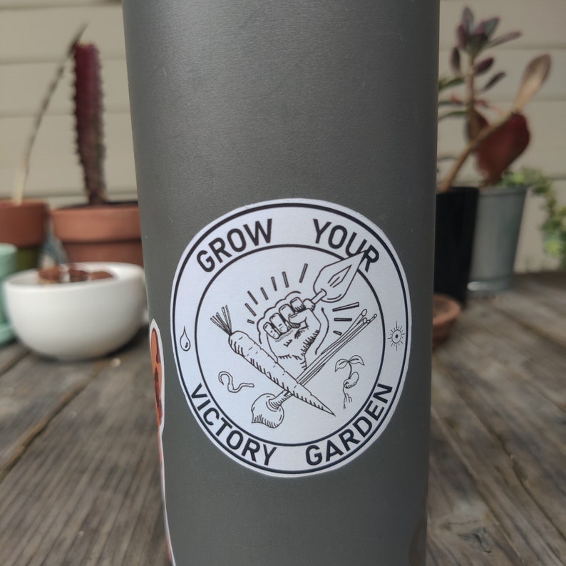 K&ouml;nnte beinhalten: Eine graue Wasserflasche mit einem wei&szlig;en Aufkleber, auf dem "Grow Your Victory Garden" steht. Der Aufkleber zeigt eine Schwarzwei&szlig;illustration einer Faust, die ein Gartenger&auml;t, eine Karotte und andere Gartenelemente h&auml;lt.