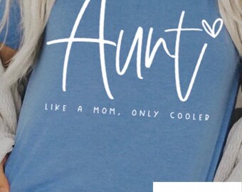 Puede incluir: Una camiseta azul claro con texto blanco que dice "Tía como una madre, solo que más cool" y un pequeño corazón. El texto está en una fuente de letra manuscrita. La camiseta tiene un logotipo blanco en la esquina inferior derecha que dice "Wilcox Creations".