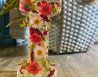 Könnte beinhalten: Ein klarer Harzbuchstabe I mit getrockneten Blumen und Blattgold eingebettet. Der Buchstabe steht auf einer Holzoberfläche.