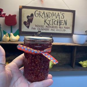 May include: A jar of homemade jam with a colorful ribbon tied around the lid. The jam is a deep red color and appears to be filled with fruit. The jar is held in a hand, with a sign in the background that says "Grandma's Kitchen."