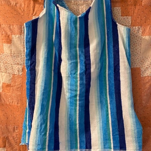 May include: A sleeveless tank top made of terry cloth fabric featuring vertical stripes in various shades of blue, turquoise, and white. The garment has a V-neckline and is displayed on a white plastic hanger, showcasing the textured material.