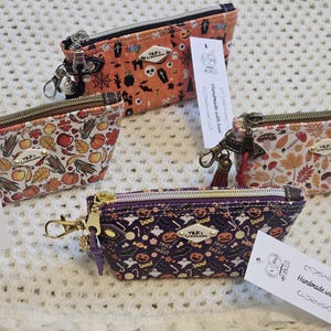 May include: Four small zippered pouches with various patterns. One is orange with Halloween motifs, another has autumn leaves, and a third features pumpkins and corn. The fourth pouch is dark purple with Halloween designs. Each has a tag that says "Handmade with love."