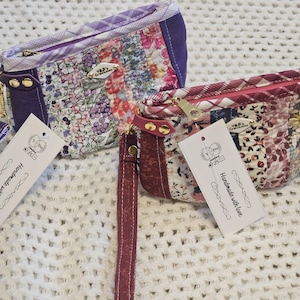 May include: Two small quilted fabric pouches with zipper closures and wrist straps. The pouches are made of patchwork fabric with floral and plaid patterns in shades of purple, pink, and white. Each pouch has a small tag attached that reads "Handmade with love".