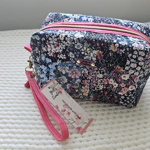 May include: A quilted cosmetic bag with a floral pattern in navy, pink, and white. The bag has a pink zipper, wrist strap, and accents. A tag with the text "YoB's Creations" is attached. The bag is on a white textured surface.