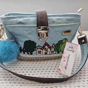 May include: A light blue quilted canvas handbag with a brown strap, a gold clasp, and a small blue pom-pom keychain. The bag features a small embroidered scene of houses and trees. The tag attached to the bag reads "Yoli's Creations Handmade with love."