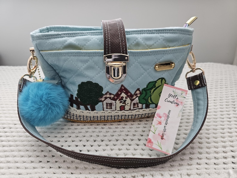 May include: A light blue quilted canvas handbag with a brown strap, a gold clasp, and a small blue pom-pom keychain. The bag features a small embroidered scene of houses and trees. The tag attached to the bag reads "Yoli's Creations Handmade with love."