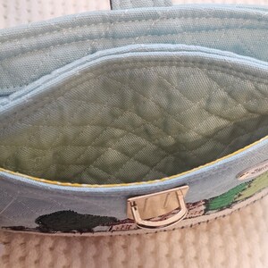 May include: A light blue quilted fabric pouch with a gold metal clasp. The pouch has a yellow trim and a green embroidered design of a house and trees. The text "Strawberry Fields" is embroidered on the pouch.