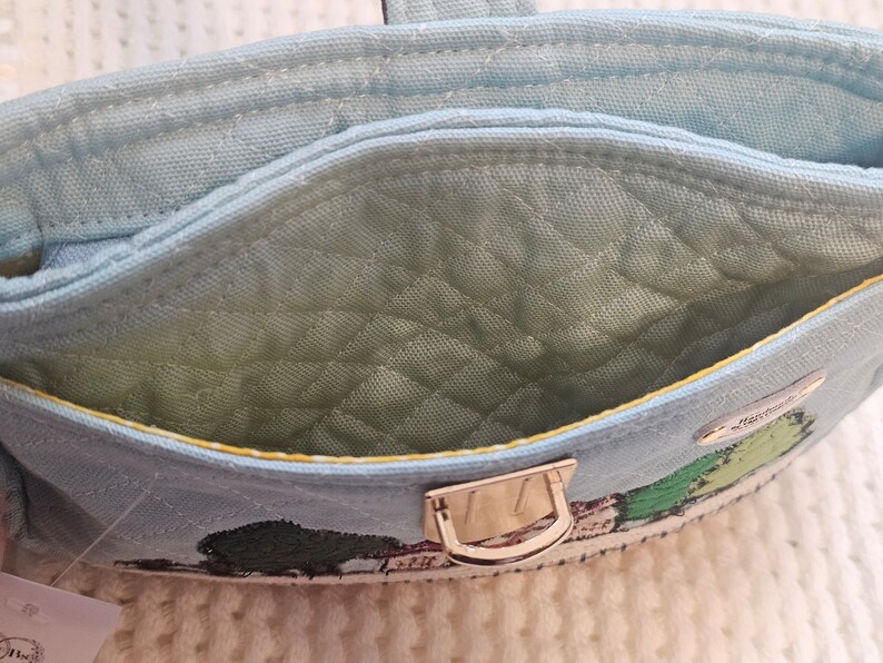 May include: A light blue quilted fabric pouch with a gold metal clasp. The pouch has a yellow trim and a green embroidered design of a house and trees. The text "Strawberry Fields" is embroidered on the pouch.