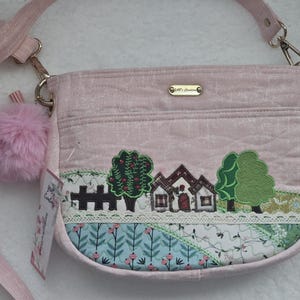 May include: A pink quilted shoulder bag with a fluffy pink pom-pom charm. The bag features an embroidered design of a small house with trees and a fence. The bag has a gold chain strap and a gold metal tag with the text "J. & J. Designs".