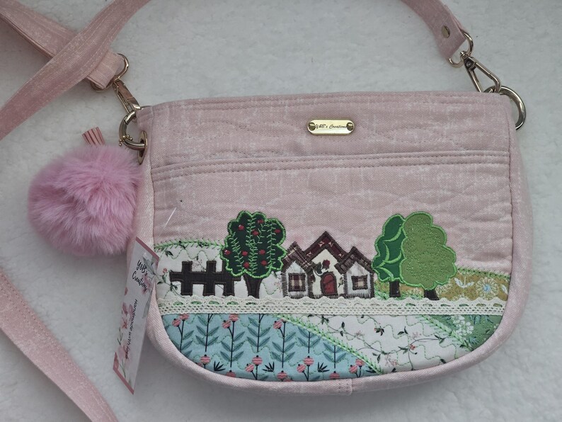 May include: A pink quilted shoulder bag with a fluffy pink pom-pom charm. The bag features an embroidered design of a small house with trees and a fence. The bag has a gold chain strap and a gold metal tag with the text "J. & J. Designs".