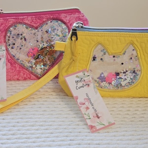 May include: Two handmade zippered pouches. One is pink with a heart-shaped window, the other is yellow with a cat-shaped window. Both have clear windows filled with glitter and small embellishments. Each has a tag that reads "Y&B's Creations, Handmade with love."