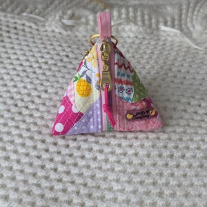 May include: A triangular fabric coin pouch featuring a patchwork design with pink, yellow, and white patterns, including a pineapple print. It has a gold-tone metal zipper, a pink pull tab, and a small brown leather tag labeled Just Creations.