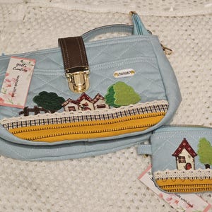 May include: A light blue quilted handbag with a gold clasp and a small matching pouch. The handbag features a whimsical design of houses, trees, and a yellow stripe. The pouch has a similar design with a house and a tree. The tag on the handbag reads "Yoli's Creations Handmade with Love."