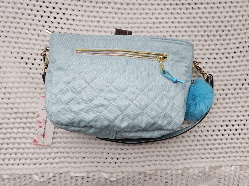 May include: A light blue quilted shoulder bag with a gold zipper and a fluffy blue charm. The bag has a black strap and a gold buckle.