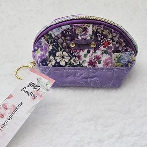 May include: A purple fabric coin purse with a floral print and a gold zipper. The purse has a small tag attached that says "Yeb's Creations Handmade with Love".