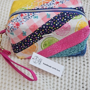May include: A colorful, quilted cosmetic bag with a zipper closure. The bag features a variety of fabric patterns, including floral, polka dot, and citrus fruit designs. It has a pink wrist strap and a tag that reads "handmade with love."
