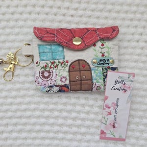 May include: A handmade, patchwork-style wallet with a house design, featuring a red roof, brown door, and floral accents. Includes a gold keychain with a blue tassel and a tag that reads "Y&B's Creations, Handmade with love."