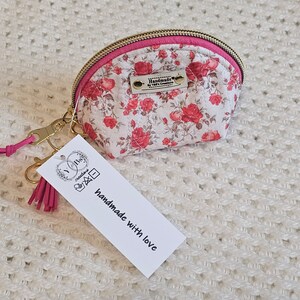 May include: A small, white fabric coin purse with a pink zipper and a floral pattern featuring red roses. The purse has a gold-colored metal key ring with a pink tassel attached. A white tag with the text "Handmade with love" is attached to the key ring.