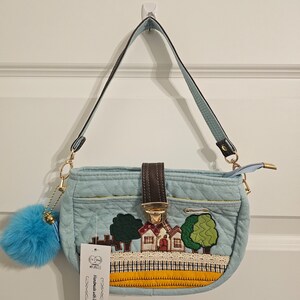May include: A light blue quilted handbag with a brown leather strap and a gold clasp. The bag features an embroidered design of a house with trees and a yellow and orange trim. The tag on the bag reads "Handmade with love".