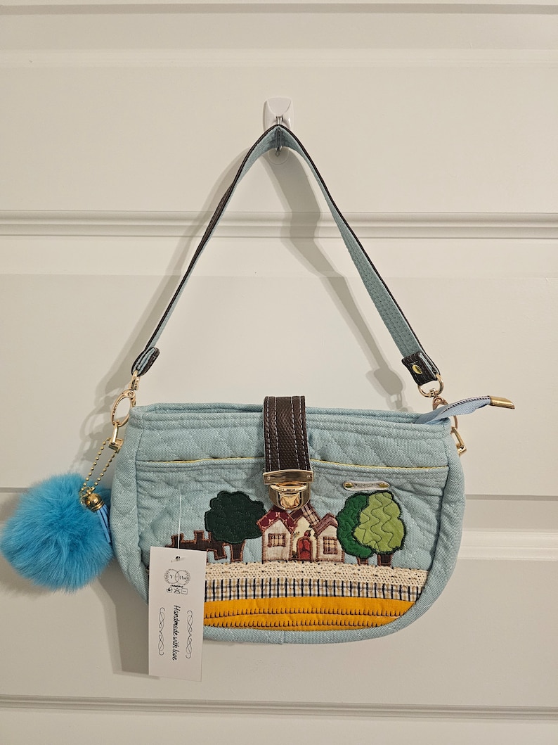 May include: A light blue quilted handbag with a brown leather strap and a gold clasp. The bag features an embroidered design of a house with trees and a yellow and orange trim. The tag on the bag reads "Handmade with love".