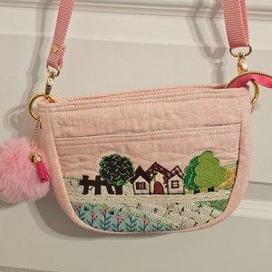 May include: A pink quilted crossbody bag with a small fluffy pink pom-pom charm. The bag features an embroidered scene of houses and trees on a green and blue background.