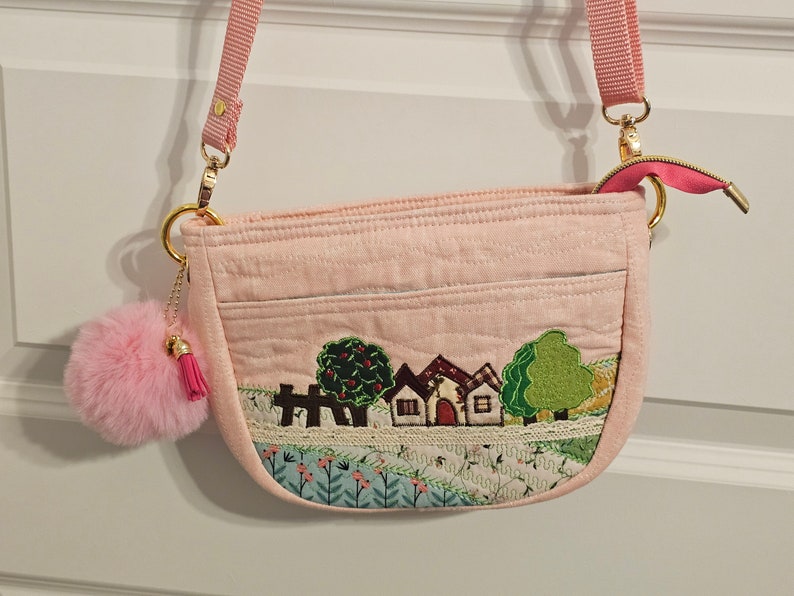 May include: A pink quilted crossbody bag with a small fluffy pink pom-pom charm. The bag features an embroidered scene of houses and trees on a green and blue background.