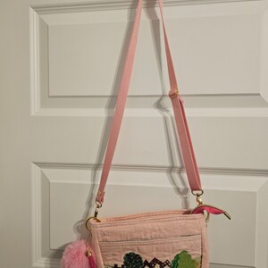 May include: A pink quilted crossbody bag with a long pink strap. The bag has a small pink pom-pom charm attached to it. The front of the bag features an embroidered design of houses and trees.