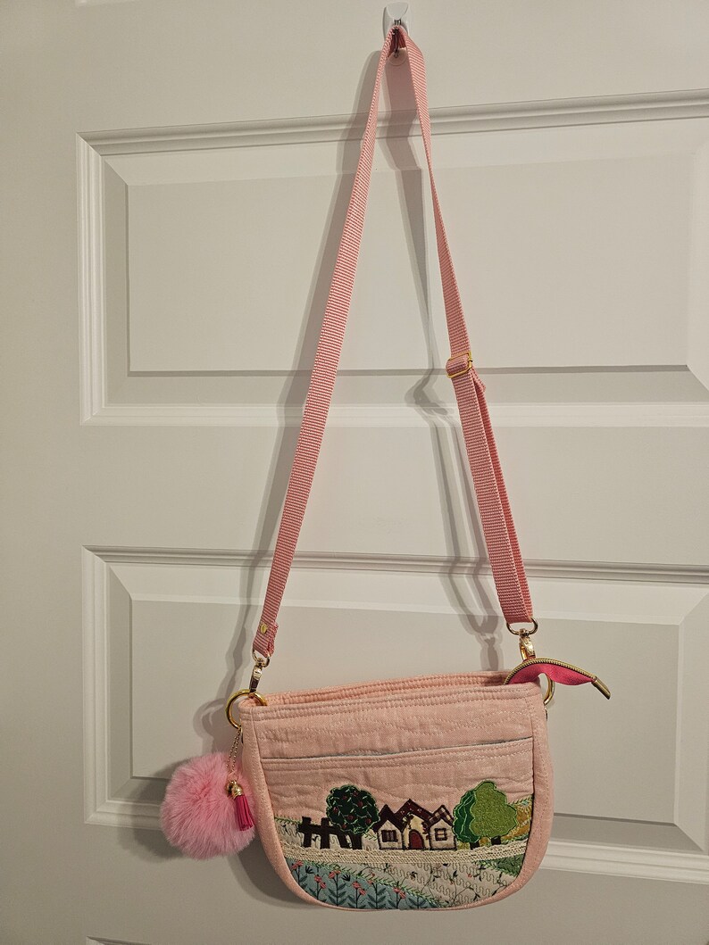 May include: A pink quilted crossbody bag with a long pink strap. The bag has a small pink pom-pom charm attached to it. The front of the bag features an embroidered design of houses and trees.