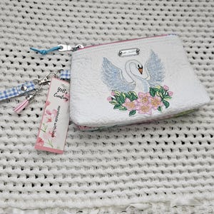 May include: White quilted zippered pouch with a white swan embroidered on it with pink flowers. The pouch has a blue and white gingham ribbon attached to it. The tag attached to the pouch reads "Yoli's Creations Handmade with Love."