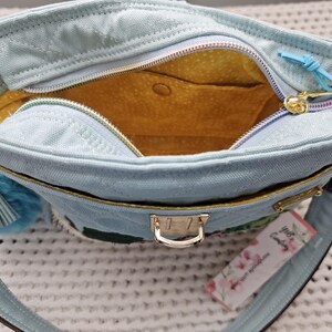 May include: A light blue canvas tote bag with a yellow lining and a gold zipper. The bag has a small pocket on the inside and a gold clasp closure. The bag has a tag that says "The Candyholic" with a floral design.