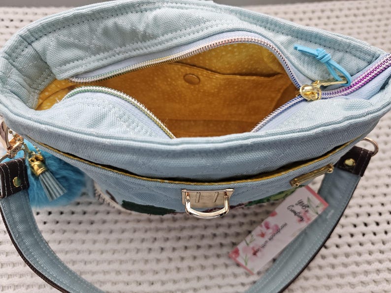 May include: A light blue canvas tote bag with a yellow lining and a gold zipper. The bag has a small pocket on the inside and a gold clasp closure. The bag has a tag that says "The Candyholic" with a floral design.