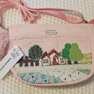 May include: A pink fabric crossbody bag with a small, fluffy pink pom-pom charm. The bag features a stitched design of a small town with houses and trees. The bag has a gold zipper and a gold chain strap. The tag on the bag reads "Handmade with love".