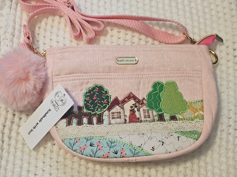 May include: A pink fabric crossbody bag with a small, fluffy pink pom-pom charm. The bag features a stitched design of a small town with houses and trees. The bag has a gold zipper and a gold chain strap. The tag on the bag reads "Handmade with love".