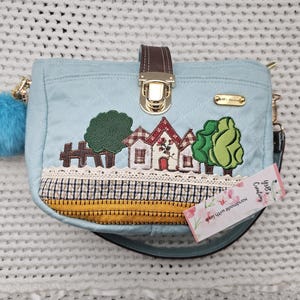 May include: A light blue quilted handbag with a gold clasp and a decorative embroidered design of houses, trees, and flowers. The bag has a short strap and a long adjustable strap. The tag on the bag reads "Handmade with love, Your's Creating".