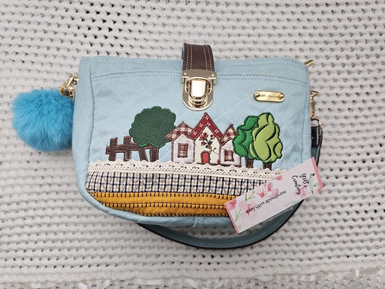 May include: A light blue quilted handbag with a gold clasp and a decorative embroidered design of houses, trees, and flowers. The bag has a short strap and a long adjustable strap. The tag on the bag reads "Handmade with love, Your's Creating".