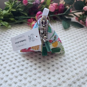 May include: A small triangular zippered pouch with a floral print and a green tassel keychain. The pouch is handmade and has a white tag with the text "Handmade with love" on it.
