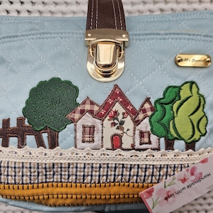 May include: A light blue canvas handbag with a brown leather handle and gold clasp. The bag features a patchwork design with embroidered houses, trees, and a fence. The bag has a white lace trim and a yellow and brown plaid fabric lining. The bag has a tag that says "Ybbs & Creativity Handmade with love".